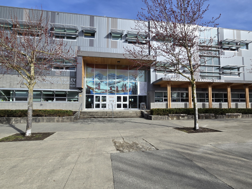 A grey building is lined by trees. 