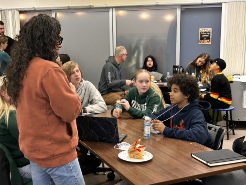 A group of a students sit around a desk while an adult stands listening to them. 