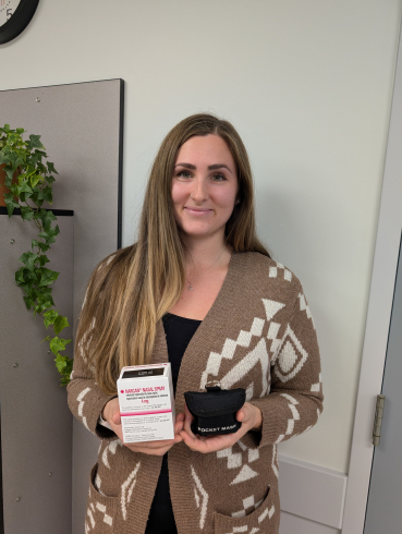 A smiling woman holds a small white box that reads "Narcan Nasal Spray" and a small black pouch that reads "pocket mask."
