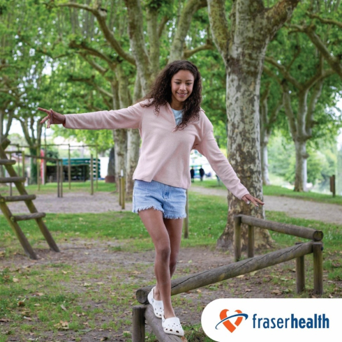 A girl with long curly brown hair balances on a log. She is in a park with leafy green trees in the background.