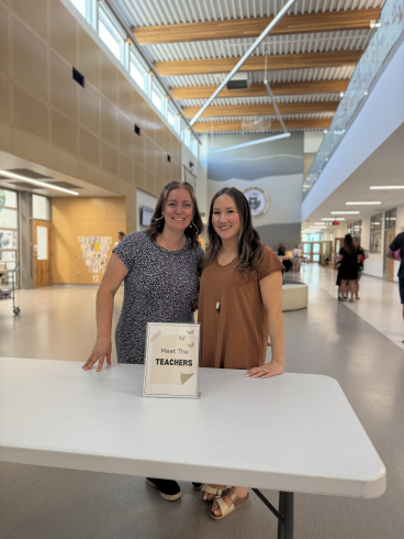Two teachers smile at the camera. On a white table in front of them a sign reads "Meet the teachers". 