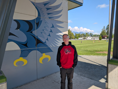 A man in a red and black hoodie stands in front of a building with a mural. Children play on a field in the background. 