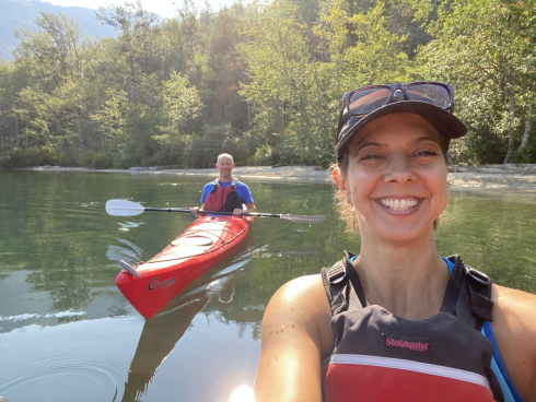 A woman in a life jacket sits in a kayak in the water. She is wearing and hat and has a big smile. There is a man in a kayak behind her who is also smiling.