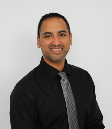 A man with a black button up shirt and tie. He is smiling. 