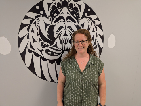 A woman in a green button up shirt stands in front of a beige wall with a circular black and white image. 