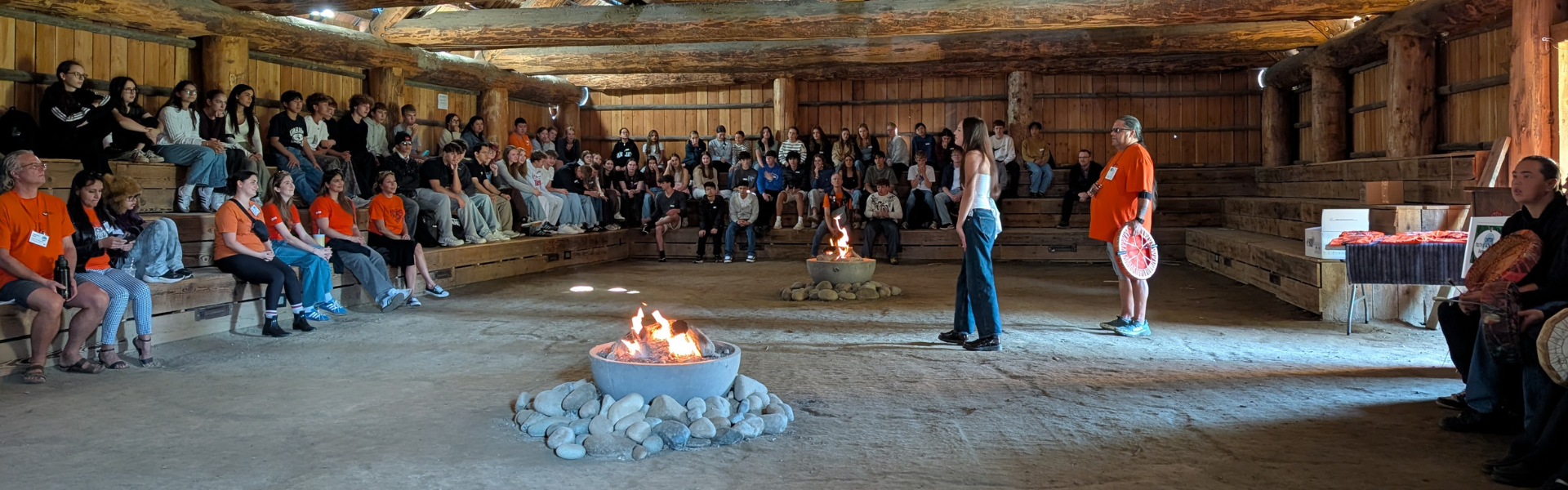 Students are gathered in a long building with great big wood beams. The walls are open on one side and a fire is burning in the middle of the dirt floor. 