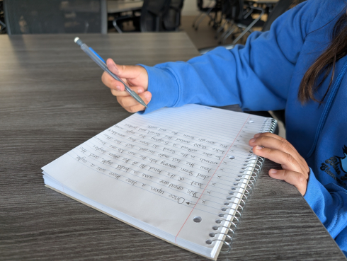 A student's hands and arms are visible as they write with a pencil in a white notebook.