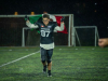 A student in football gear runs down a field holding a green, white and red flag above their head. 