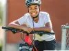 A Black youth in a white shirt rests on a bright red bike. He is wearing a helmet. 