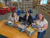 Three women sit at a table behind piles of books. They are smiling at the camera. 