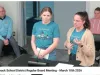 Two women in blue shirts stand at a podium. One wears a brown cardigan. They are speaking into a microphone. 
