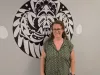 A woman in a green button up shirt stands in front of a beige wall with a circular black and white image. 