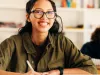 A girl with long brown hair and glasses with black frames is smiling and holding a pencil.  